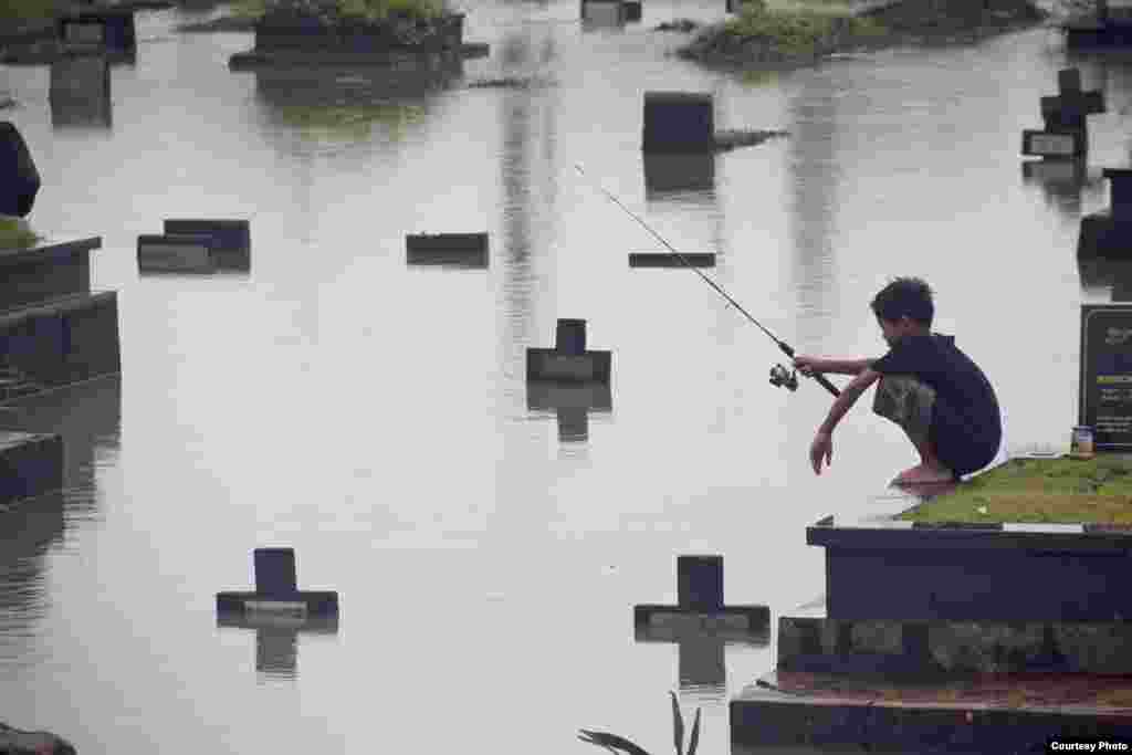 Atet Dwi Pramadia dari Indonesia meraih juara harapan lewat foto "Fishing in the Cemetery", menggambarkan anak yang memancing di sebuah tempat pemakaman yang kebanjiran di Jakarta.